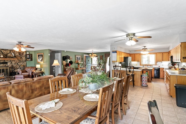 dining area featuring a stone fireplace, light tile patterned floors, and ceiling fan with notable chandelier