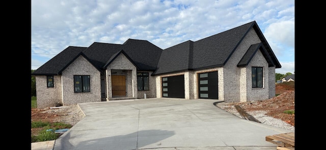 french country home with a garage, concrete driveway, roof with shingles, and brick siding