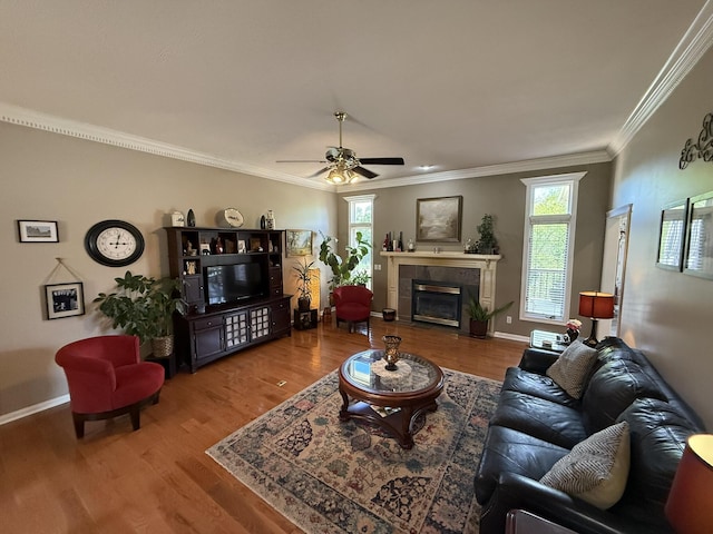 living area featuring baseboards, ornamental molding, a tiled fireplace, ceiling fan, and wood finished floors