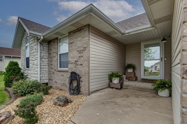 property entrance with brick siding, roof with shingles, and a patio