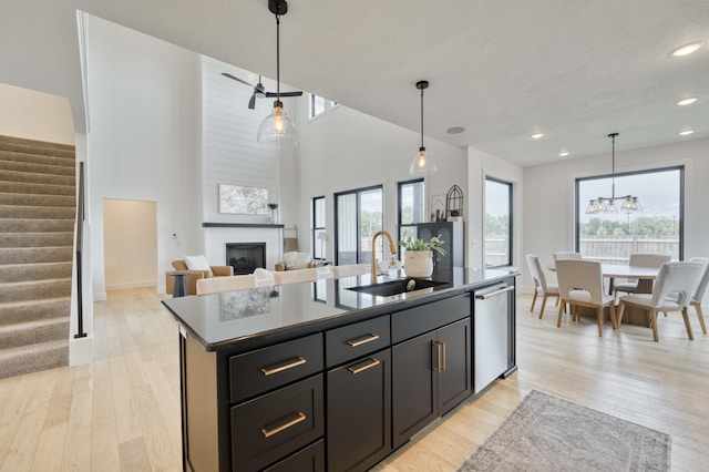 kitchen with dark countertops, stainless steel dishwasher, a sink, a center island with sink, and a large fireplace