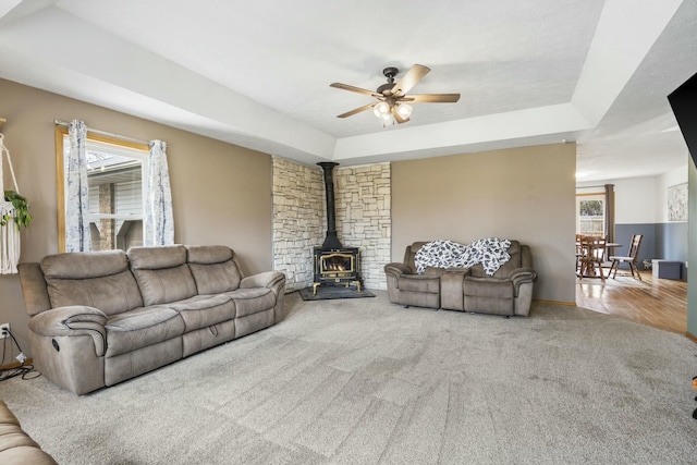 living area with a tray ceiling, a wood stove, carpet, and ceiling fan
