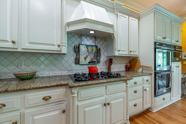 kitchen with black gas stovetop, double oven, light wood-type flooring, decorative backsplash, and custom exhaust hood