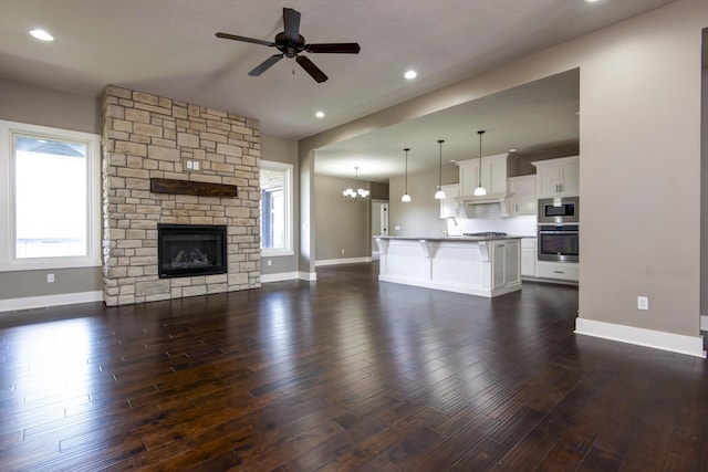 unfurnished living room featuring a stone fireplace, ceiling fan with notable chandelier, dark wood-style floors, and baseboards