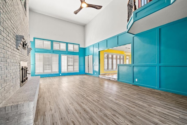 unfurnished living room featuring ceiling fan, wood finished floors, a towering ceiling, and a brick fireplace