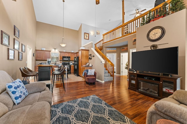 living room with a towering ceiling, light wood-type flooring, ceiling fan, stairs, and baseboards
