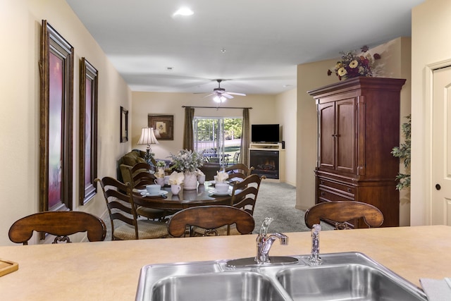 carpeted dining room featuring a sink, ceiling fan, and a warm lit fireplace