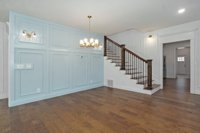 unfurnished dining area featuring dark wood-style floors, a chandelier, stairs, baseboards, and a decorative wall