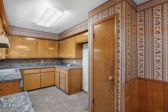 kitchen featuring a textured ceiling, a sink, under cabinet range hood, brown cabinetry, and wallpapered walls