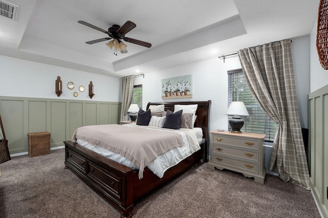 bedroom featuring a raised ceiling, multiple windows, dark colored carpet, a wainscoted wall, and ceiling fan