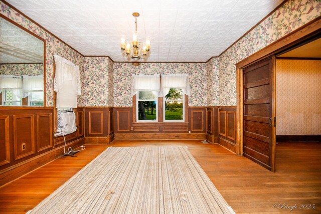 unfurnished dining area featuring wood finished floors, wallpapered walls, a chandelier, wainscoting, and an ornate ceiling