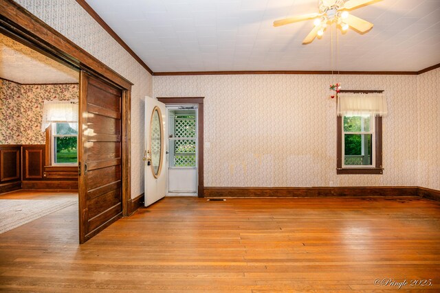 empty room with wood-type flooring, healthy amount of natural light, ceiling fan, wallpapered walls, and ornamental molding