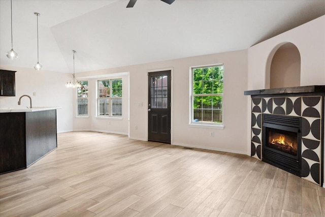 unfurnished living room featuring a tiled fireplace, light wood-style floors, ceiling fan, a chandelier, and baseboards
