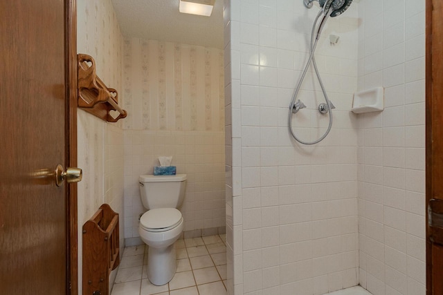 bathroom featuring a tile shower, wallpapered walls, toilet, tile patterned flooring, and a wainscoted wall