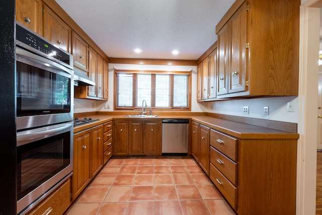 kitchen with stainless steel appliances, a sink, brown cabinetry, light tile patterned floors, and under cabinet range hood