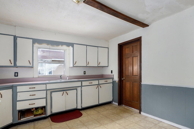 kitchen featuring a sink, light flooring, beam ceiling, white cabinets, and wainscoting