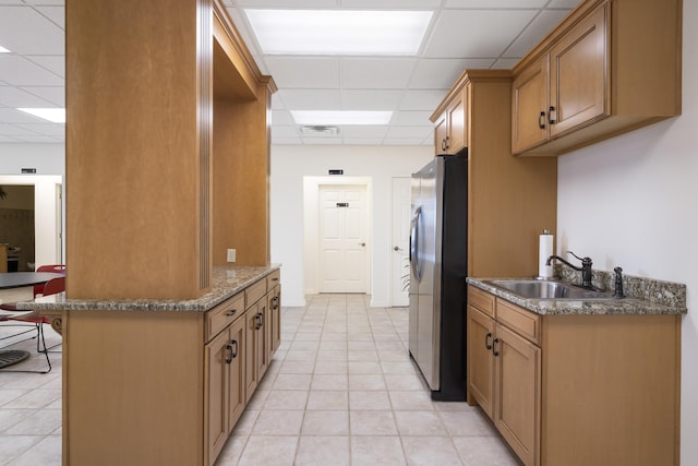kitchen with a drop ceiling, stainless steel refrigerator with ice dispenser, a sink, light tile patterned flooring, and dark stone countertops