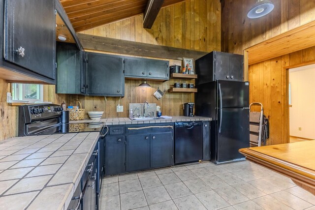 kitchen with tile counters, black appliances, wood walls, light tile patterned flooring, and a sink