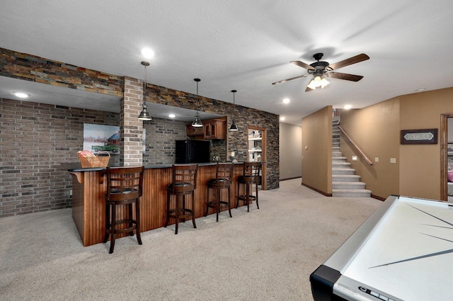 indoor dry bar with light colored carpet, a ceiling fan, freestanding refrigerator, stairs, and brick wall