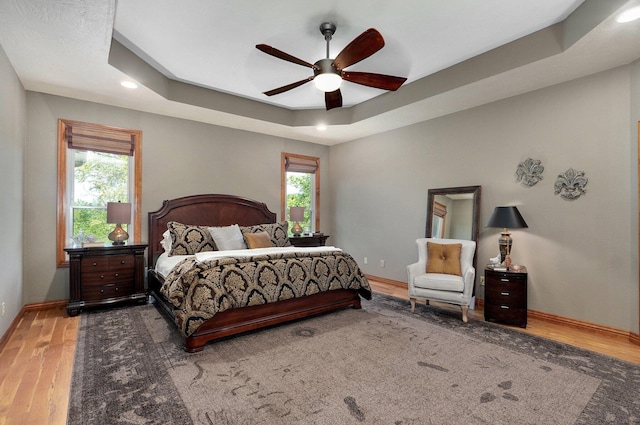bedroom featuring multiple windows, wood finished floors, baseboards, recessed lighting, and a tray ceiling