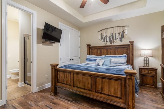 bedroom featuring a tray ceiling, wood finished floors, ensuite bath, ceiling fan, and baseboards