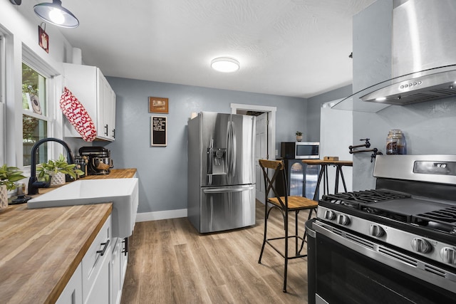 2019 N Rogers Ave, Springfield, MO 65803 - kitchen featuring appliances with stainless steel finishes, butcher block countertops, wall chimney range hood, white cabinetry, and light wood-style flooring