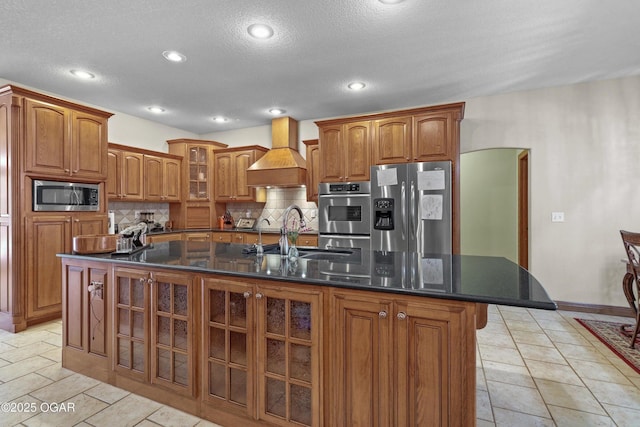 318 Fox Fire Ct, Joplin, MO 64801 - kitchen with stainless steel appliances, premium range hood, tasteful backsplash, a center island with sink, and brown cabinets