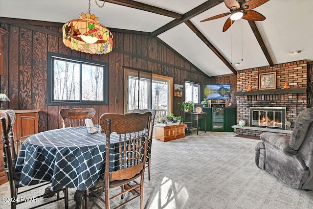 dining space featuring light carpet, ceiling fan, wooden walls, a fireplace, and vaulted ceiling with beams