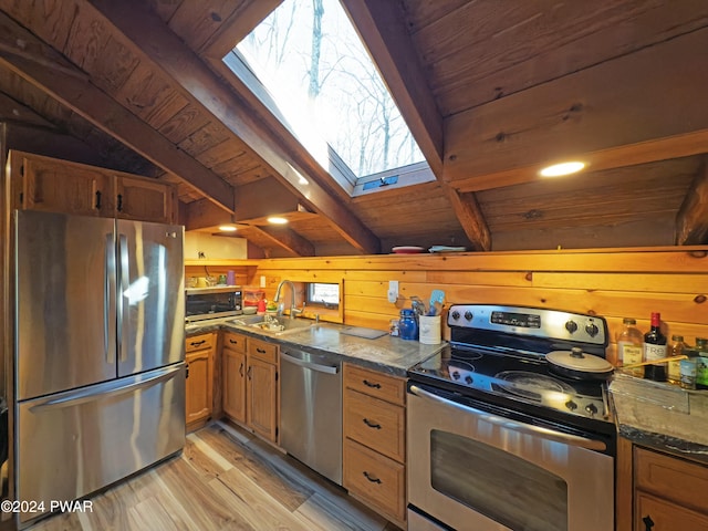 kitchen with wood walls, sink, vaulted ceiling with skylight, appliances with stainless steel finishes, and light hardwood / wood-style floors