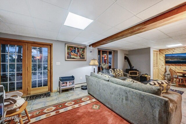 tiled living room featuring a drop ceiling, wooden walls, baseboard heating, and a wood stove