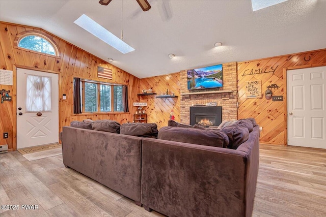 living room with wood walls, ceiling fan, a brick fireplace, vaulted ceiling with skylight, and a textured ceiling