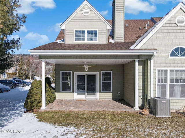 snow covered property with a shingled roof, a ceiling fan, a patio, a chimney, and cooling unit