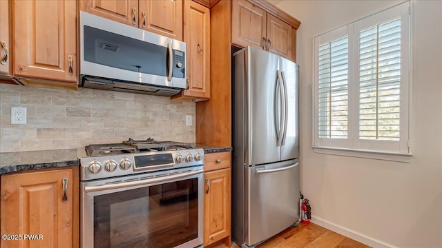 kitchen with stainless steel appliances, backsplash, brown cabinetry, dark stone counters, and baseboards