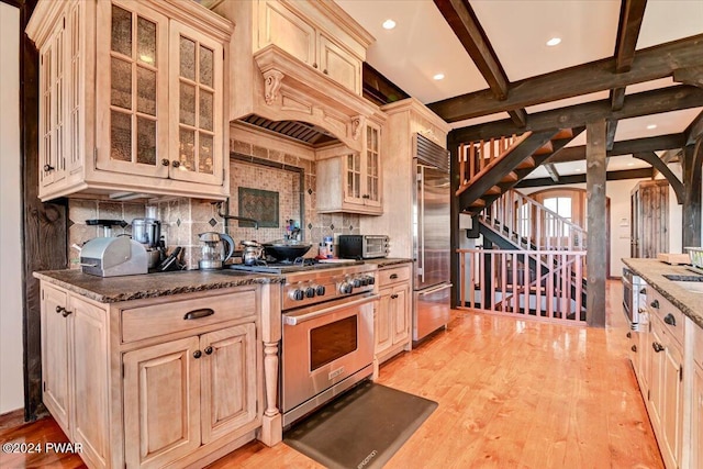 kitchen with tasteful backsplash, premium appliances, light hardwood / wood-style flooring, dark stone countertops, and beamed ceiling