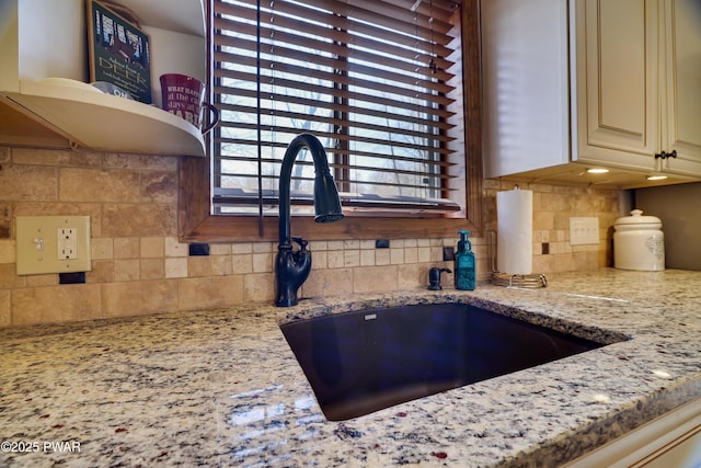 interior details with open shelves, decorative backsplash, light stone countertops, cream cabinets, and a sink
