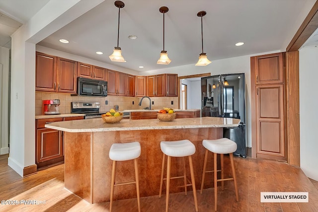 kitchen featuring brown cabinetry, an island with sink, light wood-style flooring, black appliances, and a sink