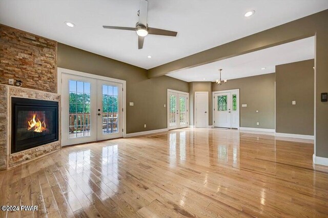 unfurnished living room with ceiling fan with notable chandelier, a stone fireplace, light hardwood / wood-style flooring, and french doors