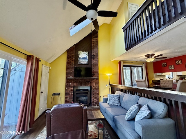 living room featuring hardwood / wood-style floors, high vaulted ceiling, a skylight, ceiling fan, and a brick fireplace