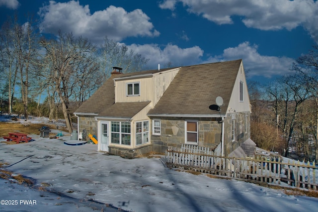 snow covered rear of property featuring stone siding, fence, and a chimney