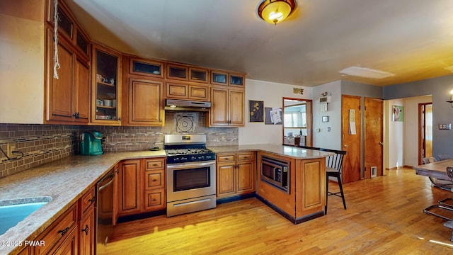 kitchen with brown cabinets, stainless steel appliances, glass insert cabinets, a peninsula, and under cabinet range hood