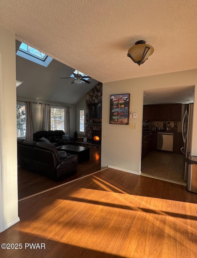 living area with vaulted ceiling with skylight, ceiling fan, wood finished floors, a textured ceiling, and a stone fireplace