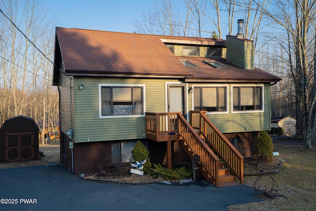 view of front facade featuring an outbuilding, a chimney, a storage unit, a shingled roof, and stairs