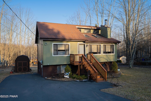 chalet / cabin featuring driveway, a storage shed, a shingled roof, a chimney, and an outbuilding