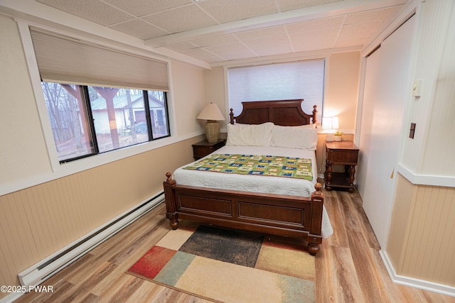 bedroom with a baseboard heating unit, wainscoting, a drop ceiling, and wood finished floors