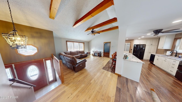 living room featuring sink, vaulted ceiling with beams, a textured ceiling, light wood-type flooring, and ceiling fan with notable chandelier