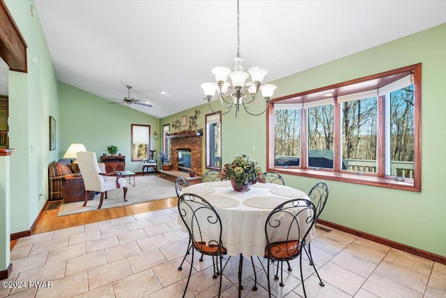 tiled dining space with ceiling fan with notable chandelier, vaulted ceiling, and a stone fireplace