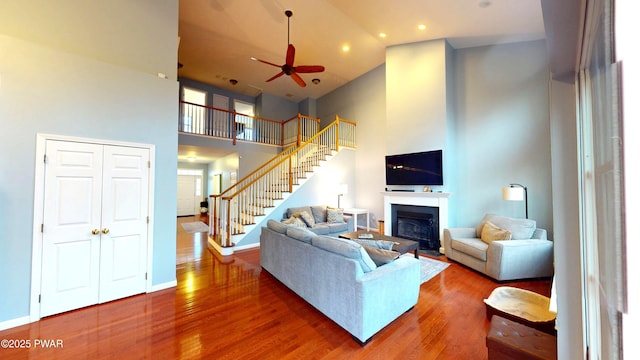 living room featuring a towering ceiling, wood finished floors, a ceiling fan, a fireplace, and stairs