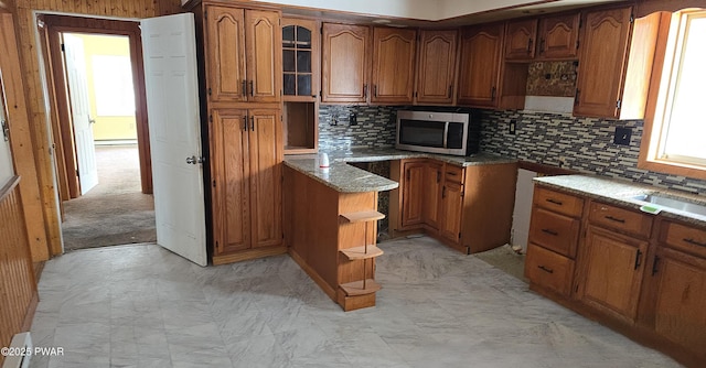 kitchen featuring brown cabinets, tasteful backsplash, stainless steel microwave, glass insert cabinets, and light stone countertops