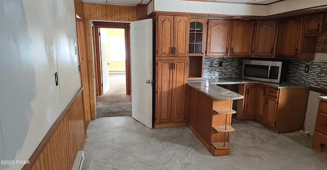 kitchen featuring light stone counters, brown cabinets, stainless steel microwave, glass insert cabinets, and wood walls