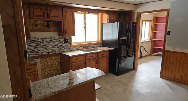 kitchen with light stone counters, wooden walls, black fridge, wainscoting, and brown cabinetry